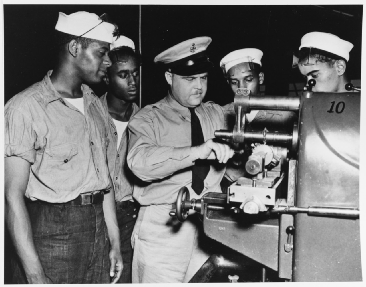 Three Black men in naval work uniforms watch a white man in khaki uniform working on machinery.