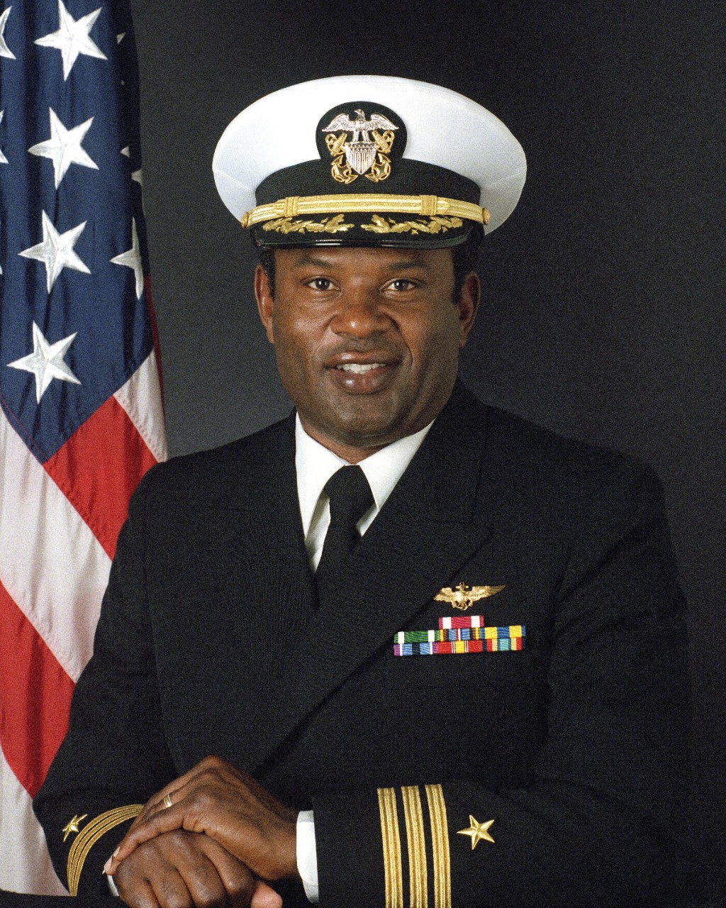 Portrait of an African-American man in Navy blue dress uniform, with the American flag displayed in background.