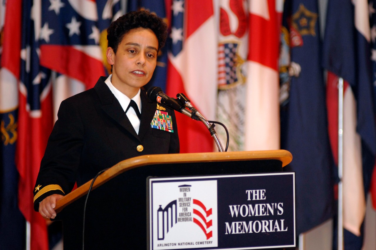 An African American woman in Navy dress blue uniform stands behind a podium with a sign for the Women's Memorial. In background, there is a display of algs.
