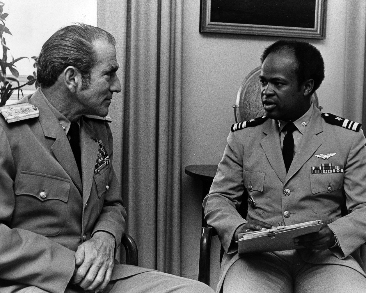 A white man in khaki Navy uniform sits facing an African American man in khaki Navy uniform.