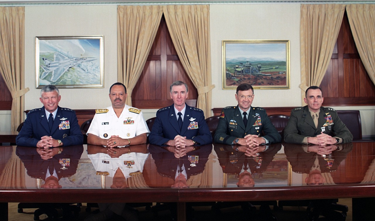 Five men sit in a row at a table in a conference room, all are in military uniform.