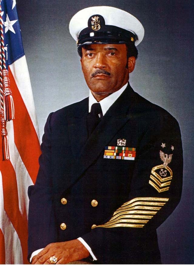 Photograph of an African American man in formal Navy blue uniform with the American flag displayed in the background on the left.