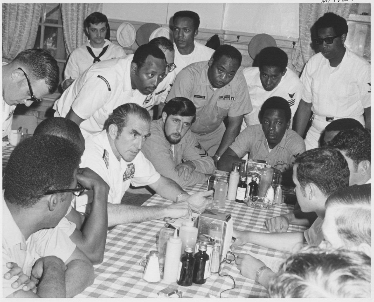 A White man in a white Navy uniform sits in the middle of a group of men, both White and Black, at a table. 