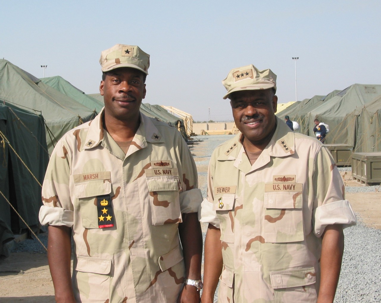 Two Black officers, both with cover, in desert khaki uniforms with tents in the background.