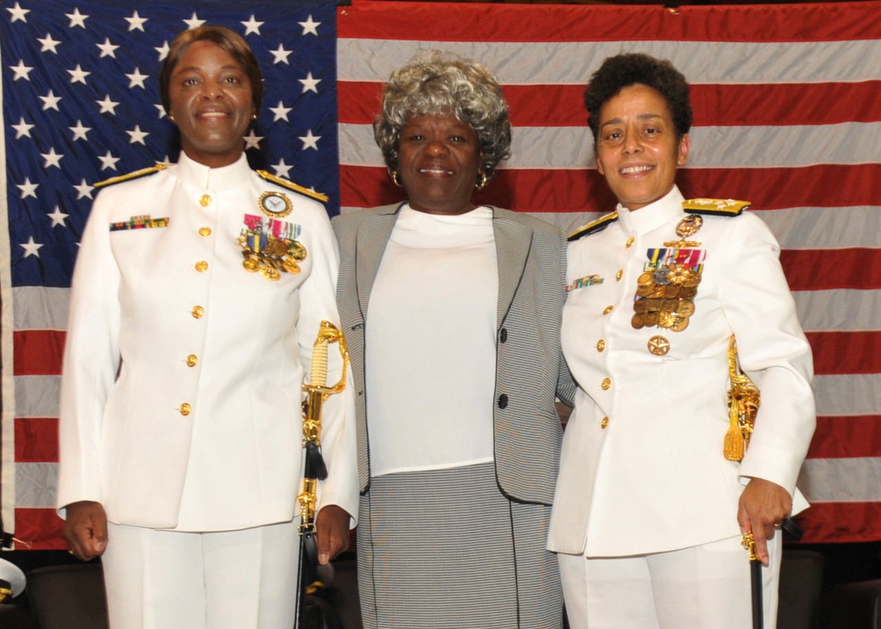 Thee African American women stand together in a group in front of the American flag, the women to the left and the right are in the formal Navy white uniform.