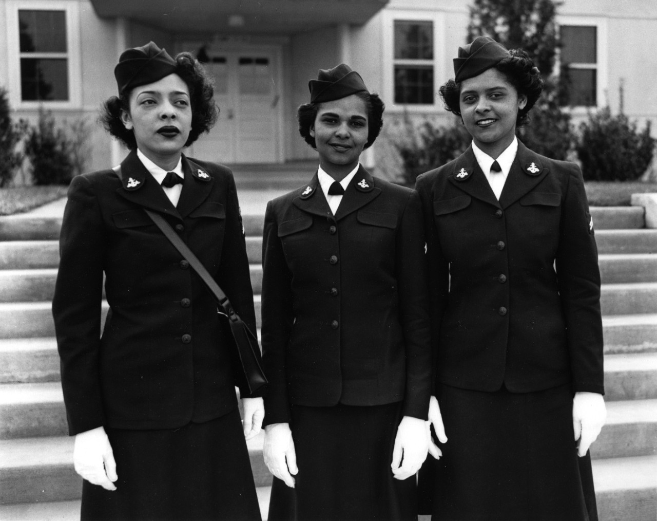 Three African American women in Navy uniform standing in a row in front of the exterior of a building with stairs. 