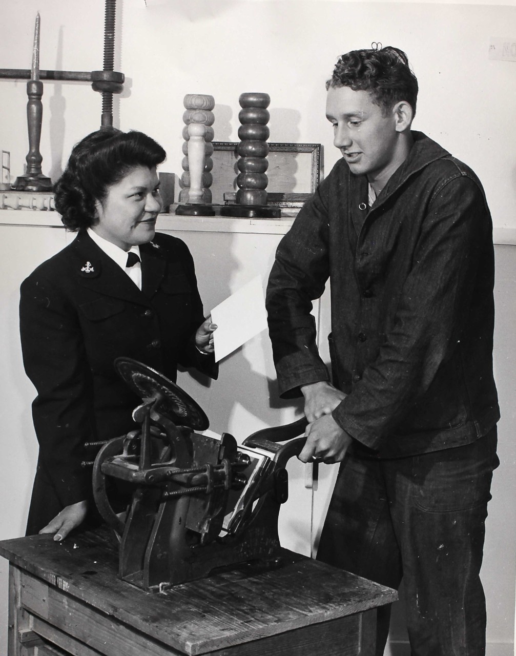 Woman in uniform assist a seaman in uniform with the use of a printing press. 