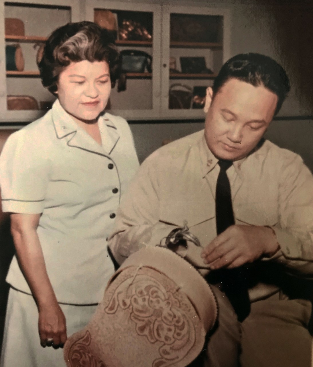 Woman in uniform looks over the shoulder of a man in uniform working on a leather craft project. 