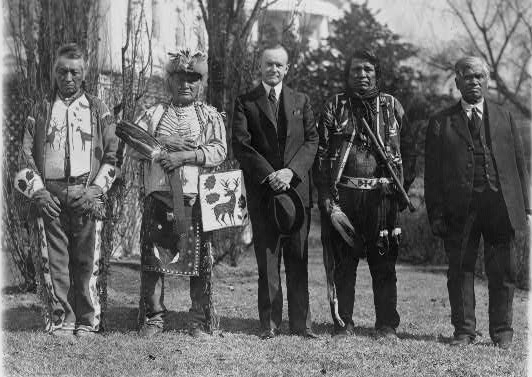 Group portrait of four Native American men, including three in tribal attire, with a White man in a suit, all standing outside.