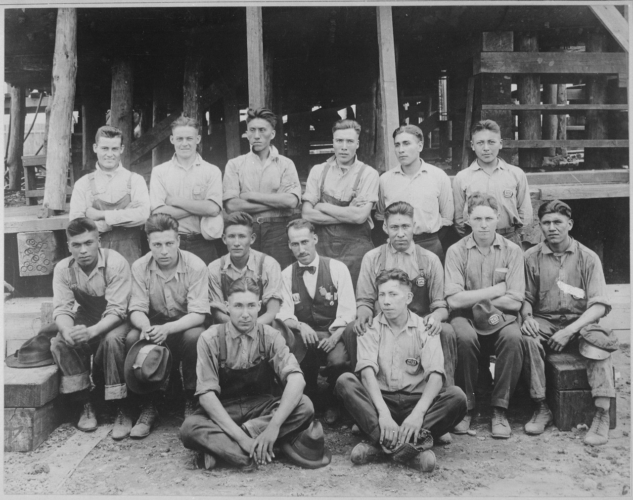 Twenty-five young men in three rows, first row is seated, in an industrial yard