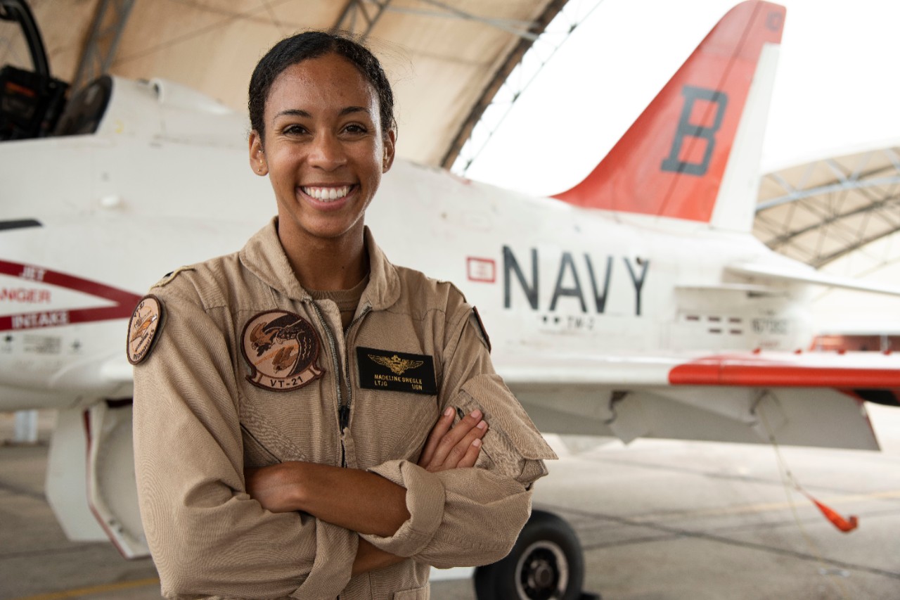 A woman in a flight suit stands with arms crossed in front of an aircraft.