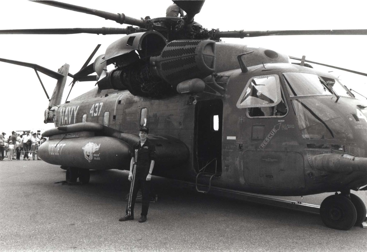 A woman stands in a flight suit in front of a helicopter. 