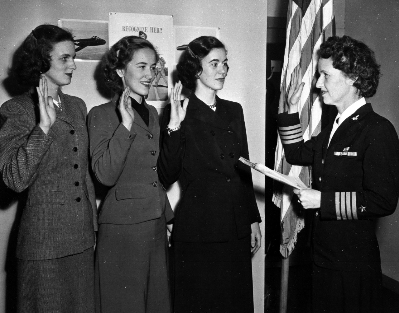 Three women in civilian clothing hold up their right hands facing a woman in uniform.