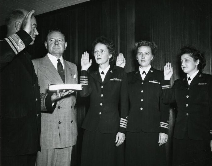 Three women in uniform raise their right hands facing a man in uniform with a main in a suit in attendance. 