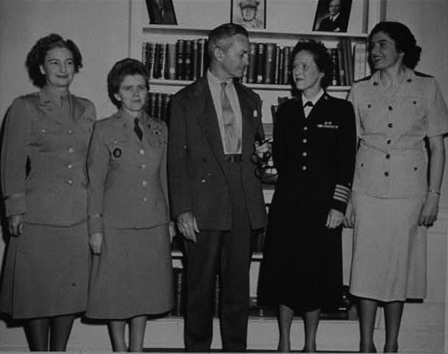 Five individuals, four women and one man, stand in a row in front of a bookcase. 