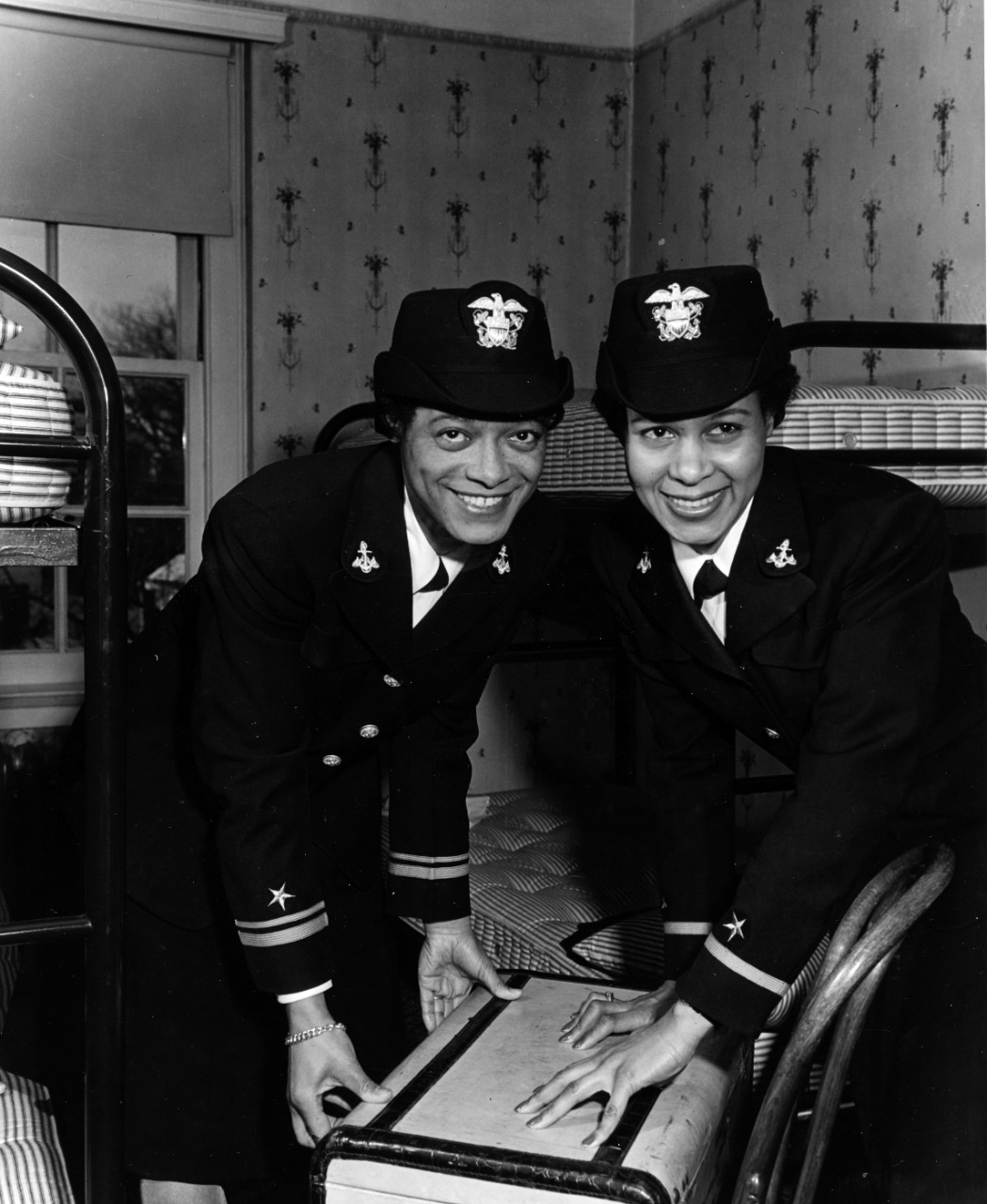 Two women in uniform learning over a suitcase in a dorm room.