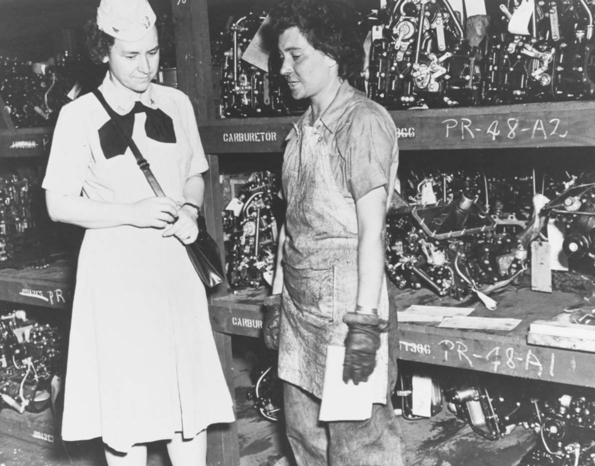 A woman in a clean dress uniform talks to a young woman wearing work gloves and an apron over grease-smudged clothes.