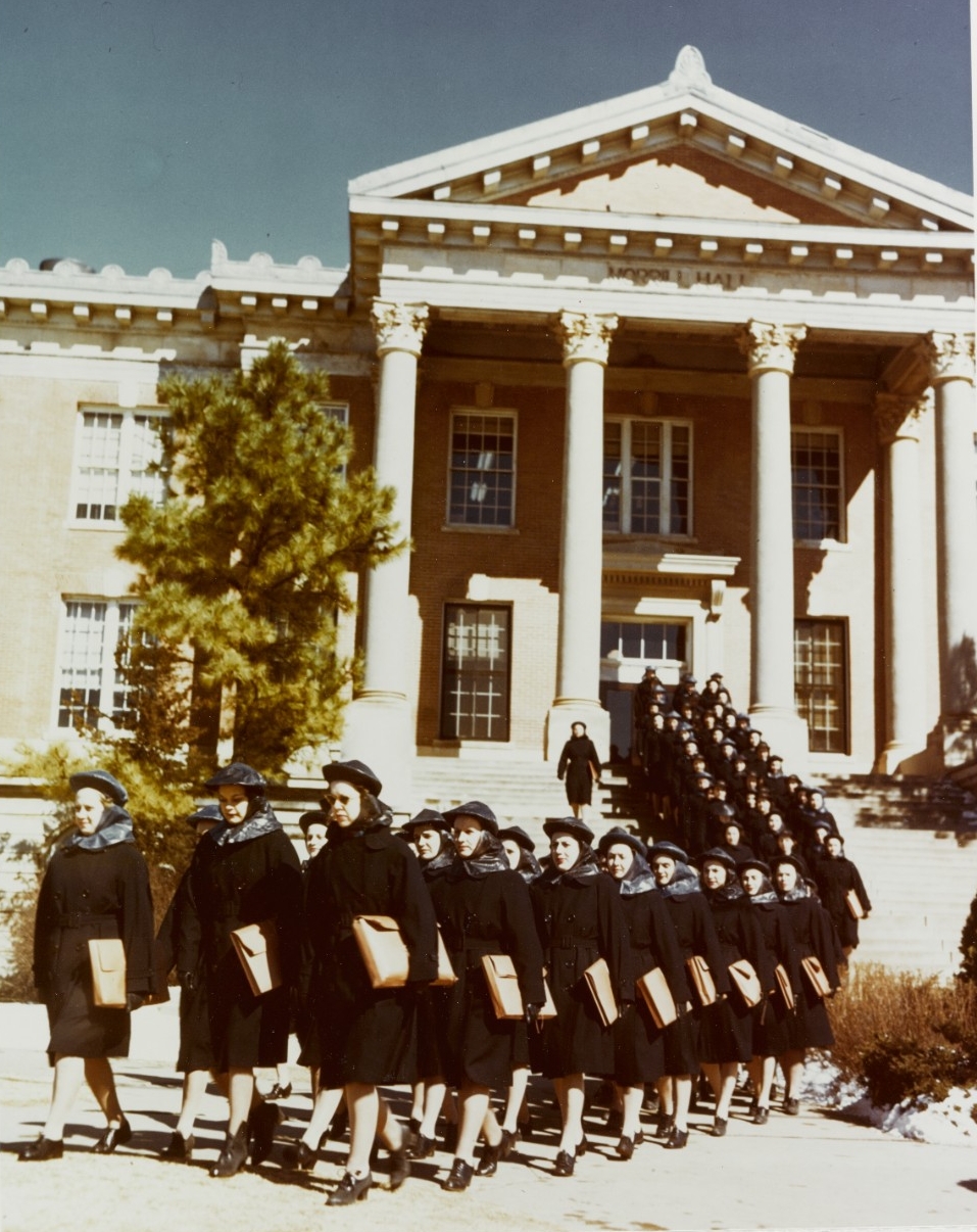 Women in black raincoats file down the steps of a large building.