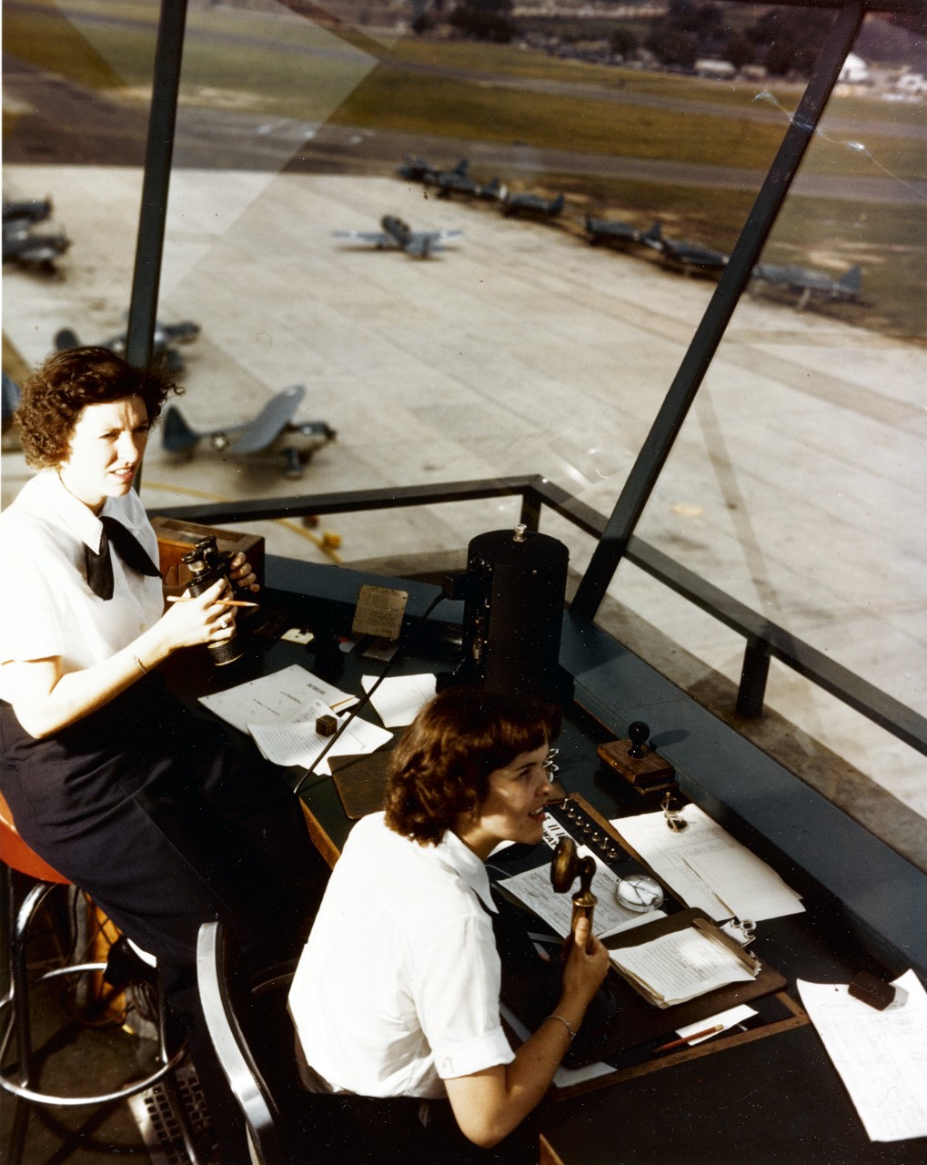 Two women in uniform sit in a control tower overlooking a runway. One wears binoculars and one is speaking into a phone. 