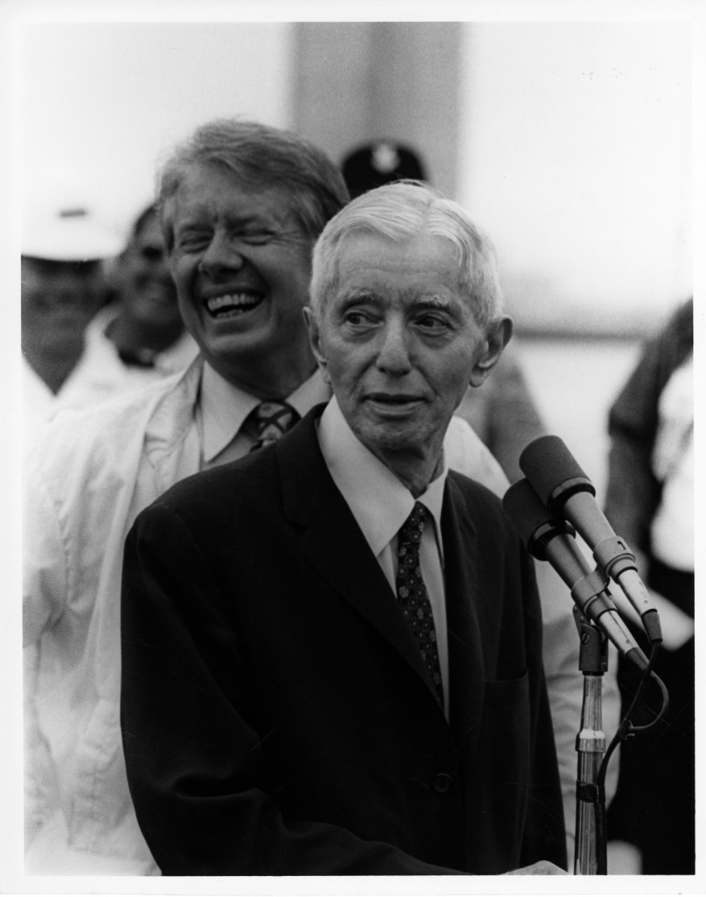 black and white photograph of a man at a microphone with another man laughing behind him