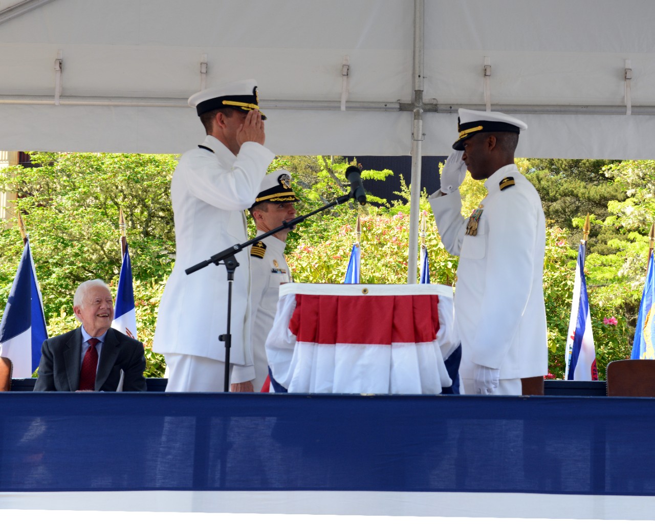 Two men in Navy uniforms salute each other while President Carter looks on from his seat.