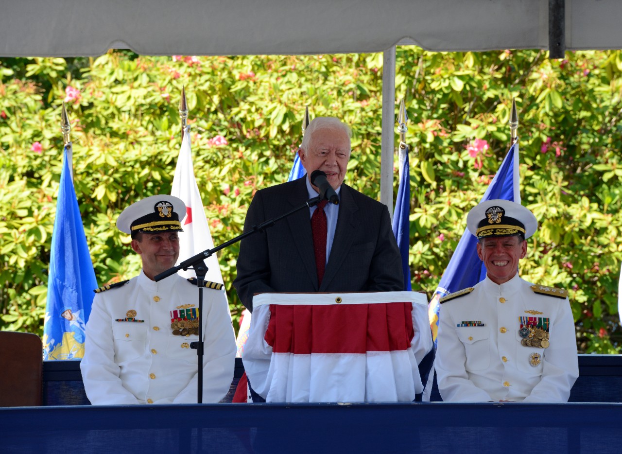 President Carter speaking at a podium with two Navy officers sitting behind him smiling. 