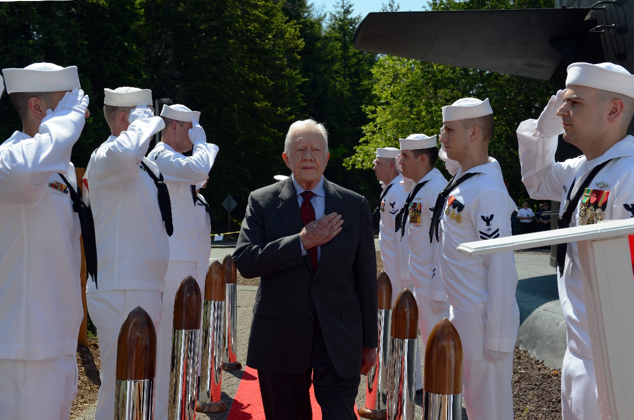 color photo of President Carter walking through a line of saluting sailors with his hand on his heart