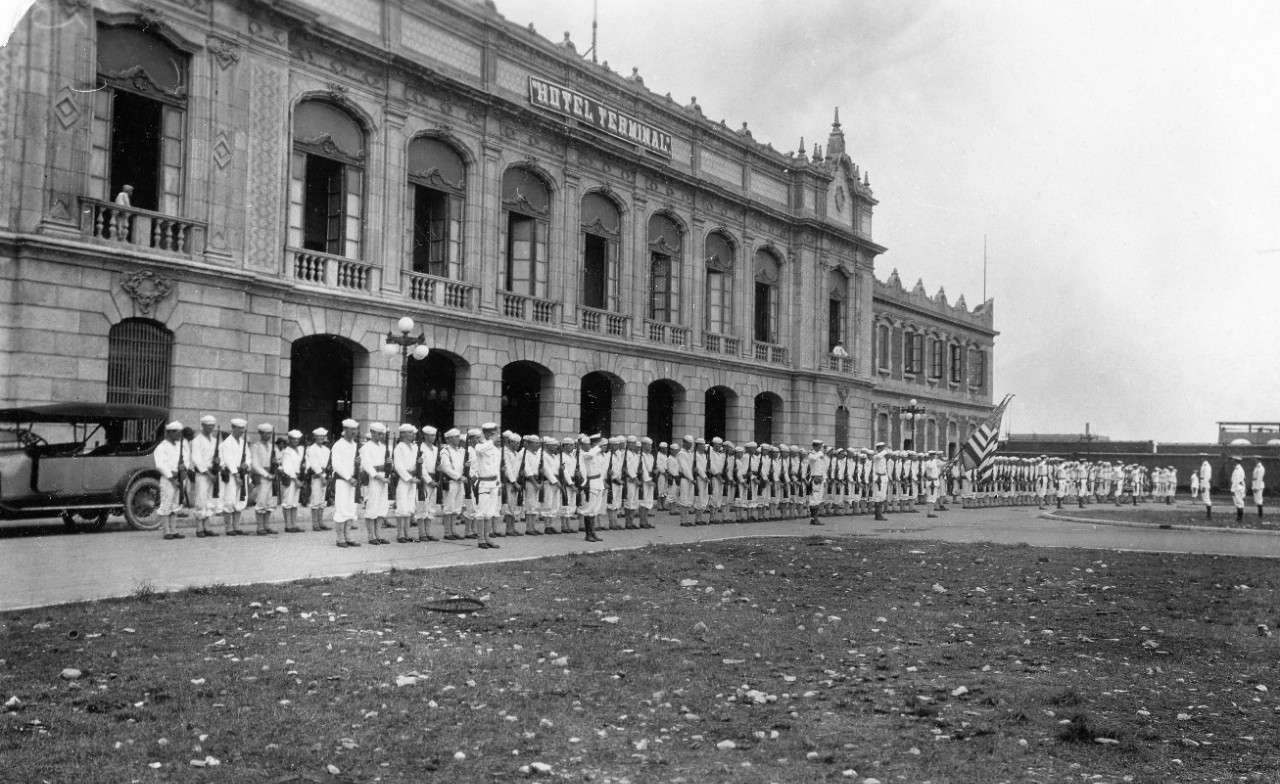Rows of white-uniformed sailors holding rifles stand in front of a stately hotel.