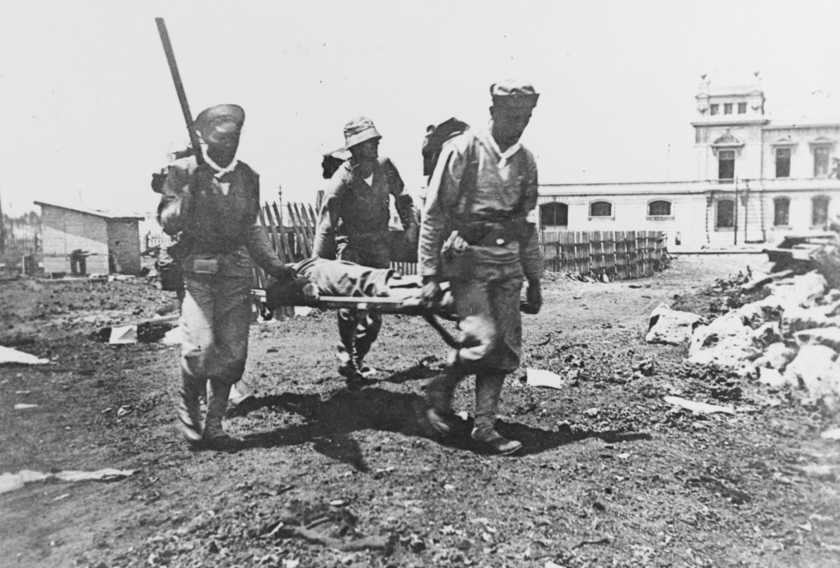 Three sailors carry a wounded man on a stretcher. Clicking on this image directs to a page to download the image.