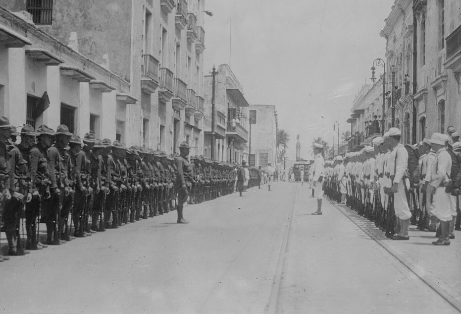 Soldiers in dark uniforms stand in a row on one side of a city street and sailors in white uniforms stand on the other side.