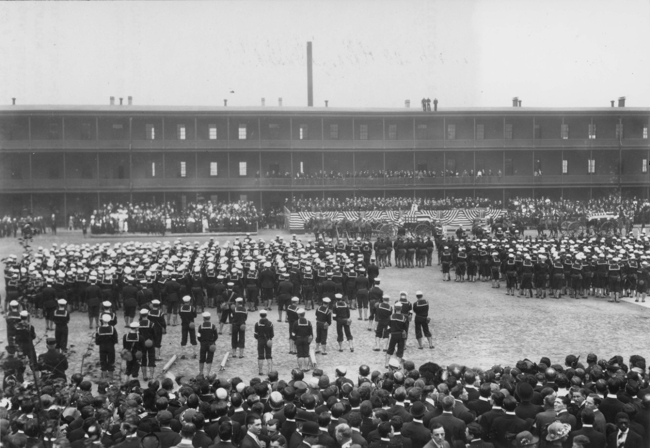 Rows of uniformed sailors stand before a crowd of civilians to watch caskets on horse-drawn caissons.