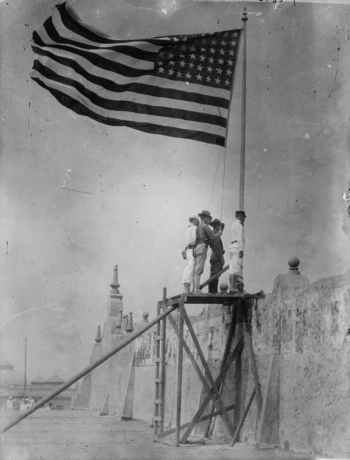 Four men stand on a wooden platform beneath an enormous American flag blowing in the wind.