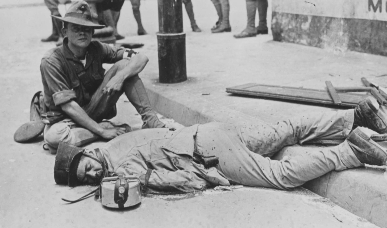 A Navy sailor sits beside a wounded Mexican soldier lying across a curb.