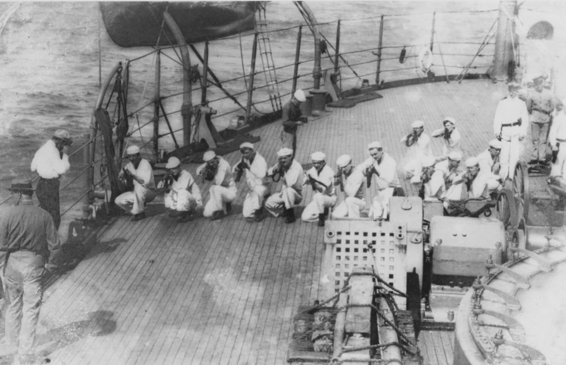 Sailors kneel and aim rifles on the deck of a ship. Clicking this image directs to a page to download the image.