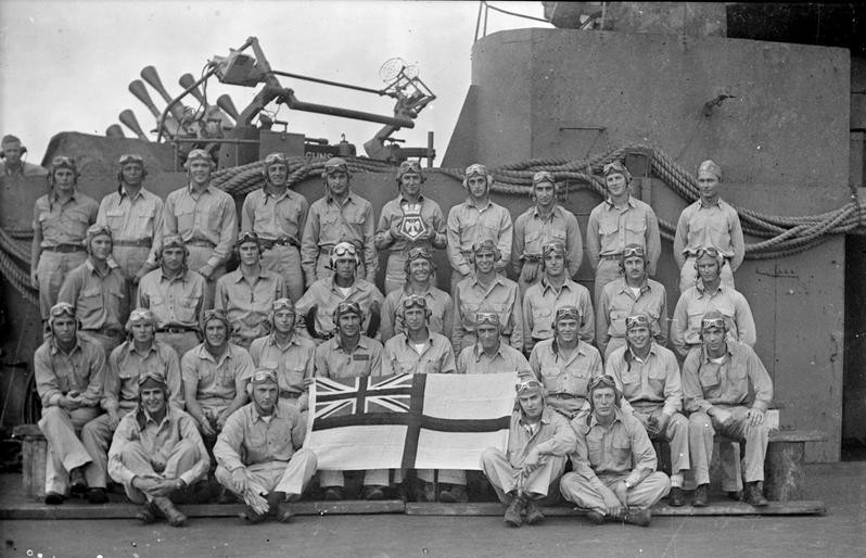 U.S. Navy VF-3 fighter pilots on board HMSVictorious pose with the British naval white ensign and the ship’s emblem, June/July 1943 