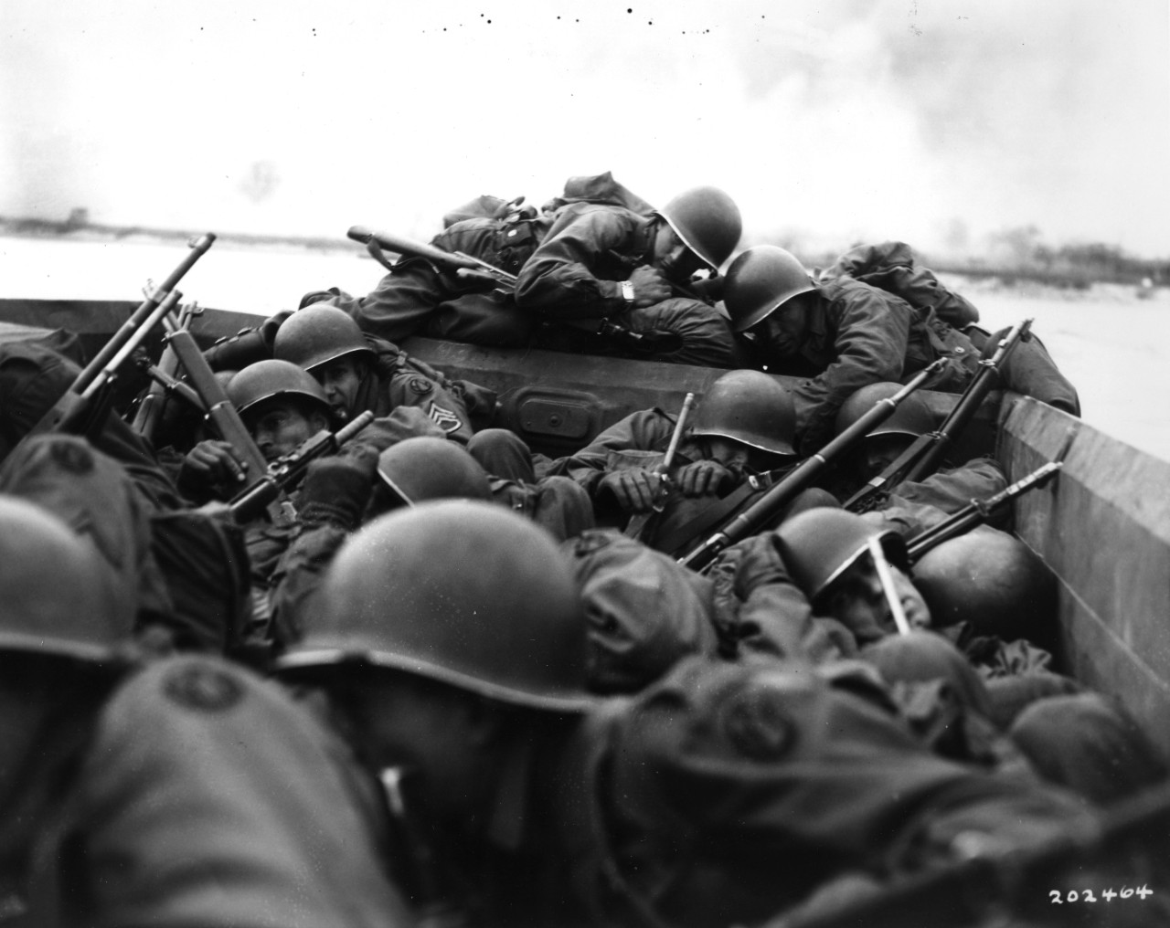 89th Division soldiers crouch low in their crowded assault boat, to escape enemy fire as they cross the Rhine River at Oberwesel, Germany, 26 March 1945. Note 89th Division insignia on men's shoulders, and variety of weapons (including M-1 rifles...