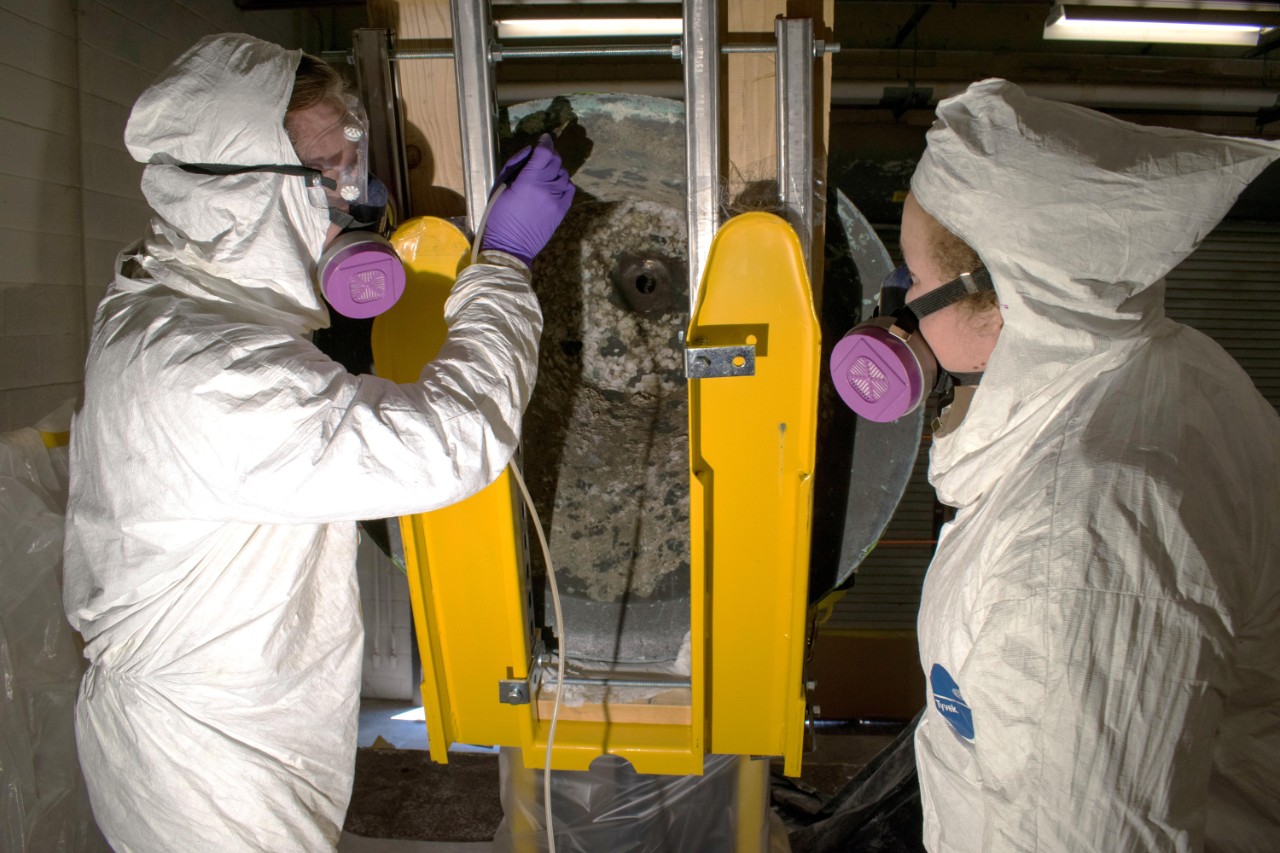 Melissa Swanson and Karl Knauer, conservators at Naval History and Heritage Command, conduct micro abrasive cleaning treatments on the underside of the USS Utah bell's rim. (U.S. Navy photo by Naval History and Heritage Command Collection Management Facility staff/Released)