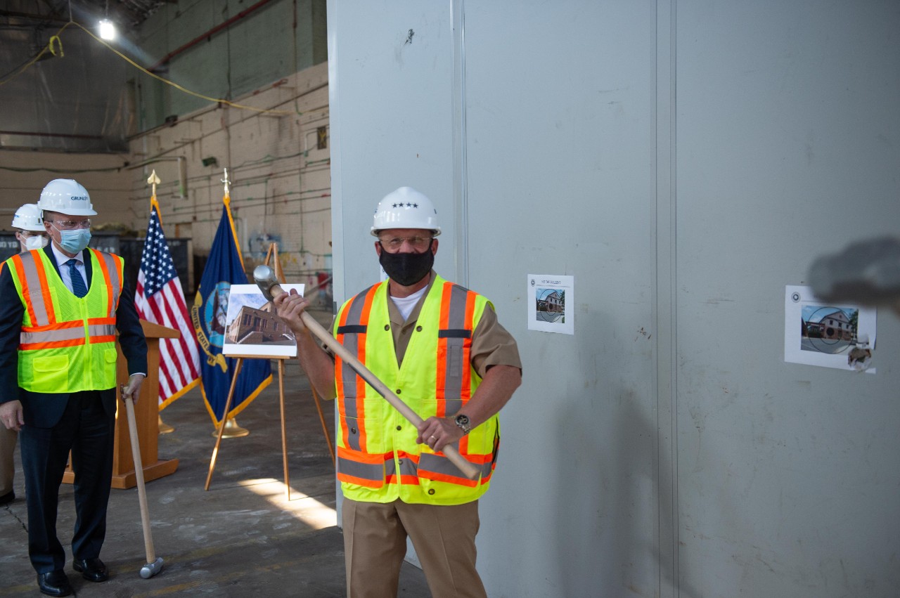 <p>CNO holding a sledge hammer in a mask, hard hat, and safety vest.</p>