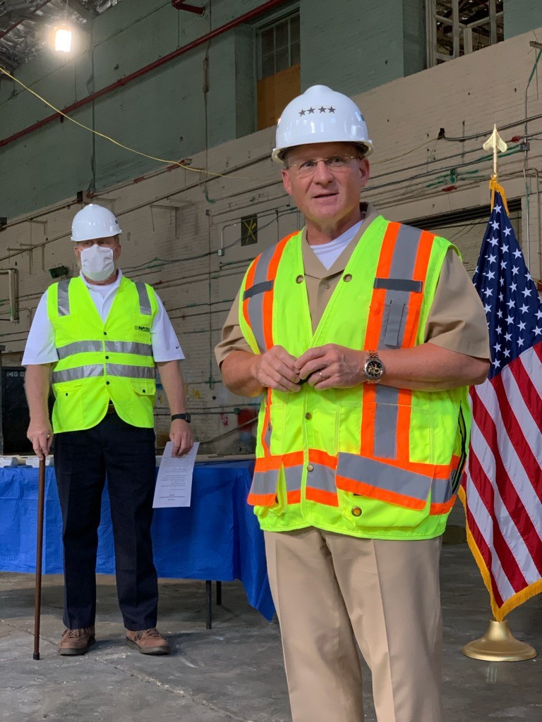 <p>CNO in hard hat with saftey vest standing in forn of an American flag.</p>