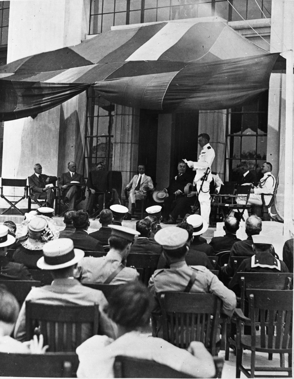 black and white photo of man in uniform on a stage under a tent in front of a seated audience.