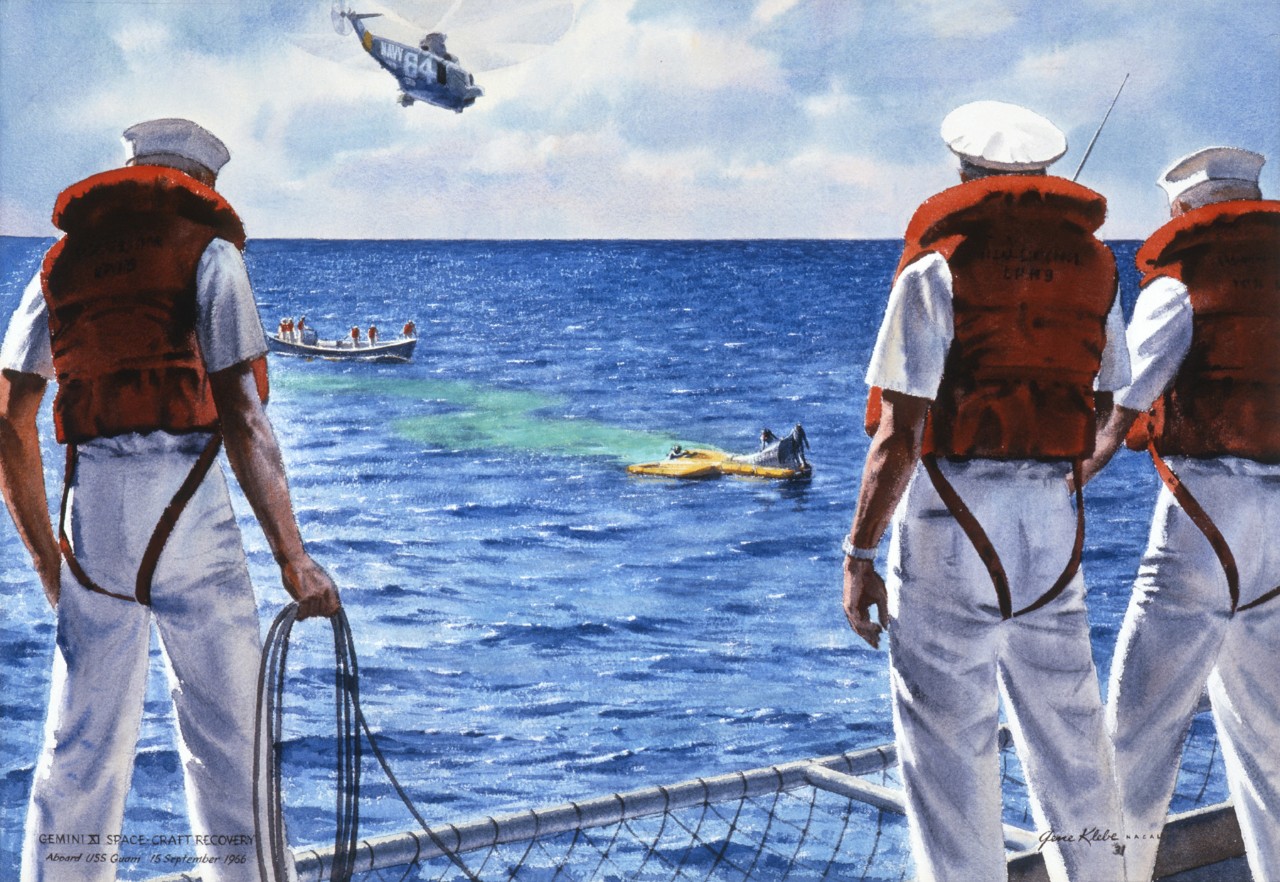 Three men stand on the deck of a ship watching the capsule in the water a helicopter fly’s overhead and a raft with divers is approaching the capsule