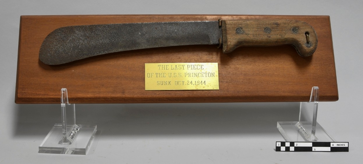 One survival non-folding machete mounted on a wood plaque. Machete has a two-piece wood grip sandwiched on the tang and a log blade. Below the machete is a brass plaque inscribed "THE LAST PIECE / OF THE U.S.S. PRINCETON / SUNK OCT. 24, 1944."