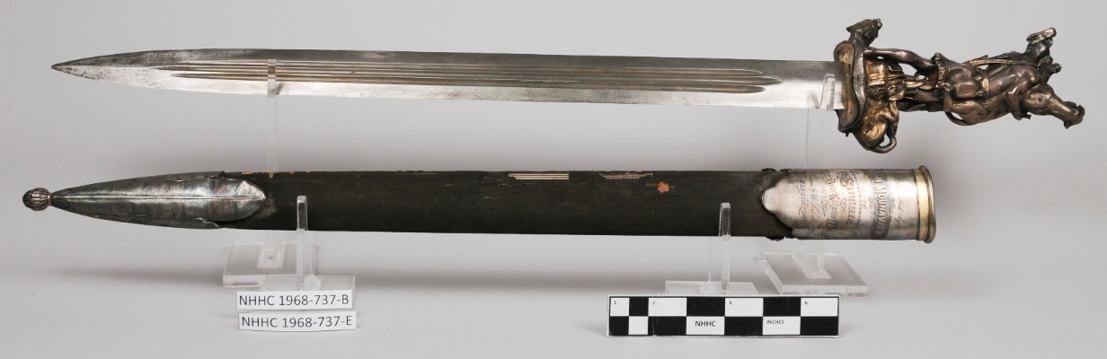 Straight-blade, double-edged sword with stylized hilt in figure of an American Indian in combat with two panthers. Scabbard is black silk with metal chape and throat. Inscription on throat.  