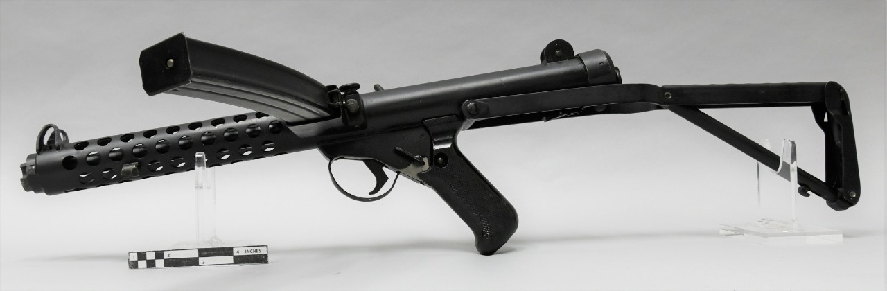Leftside of 9mm L2A3 Sterling submachine gun. Black metal finish with black plastic pistol grip. The stock is extended behind the gun. A curved metal magazine is loaded into the left side of the gun.