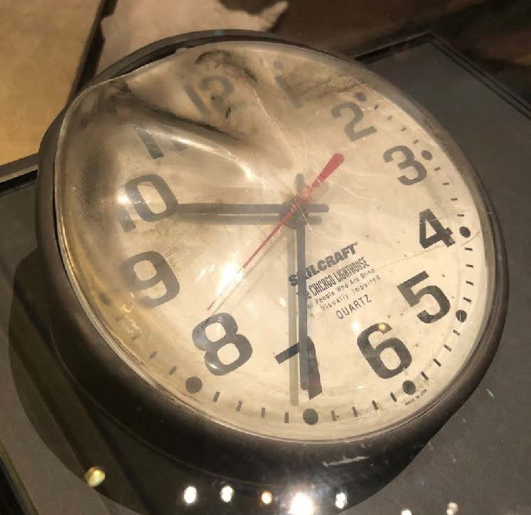 Angled view of 12-hour clock with white face and black numbers. Hands stopped at 936. Red second hand stopped at 42 seconds. Top left side of clear plastic cover blackened and warped. Recovered from Pentagon after terrorist attack.