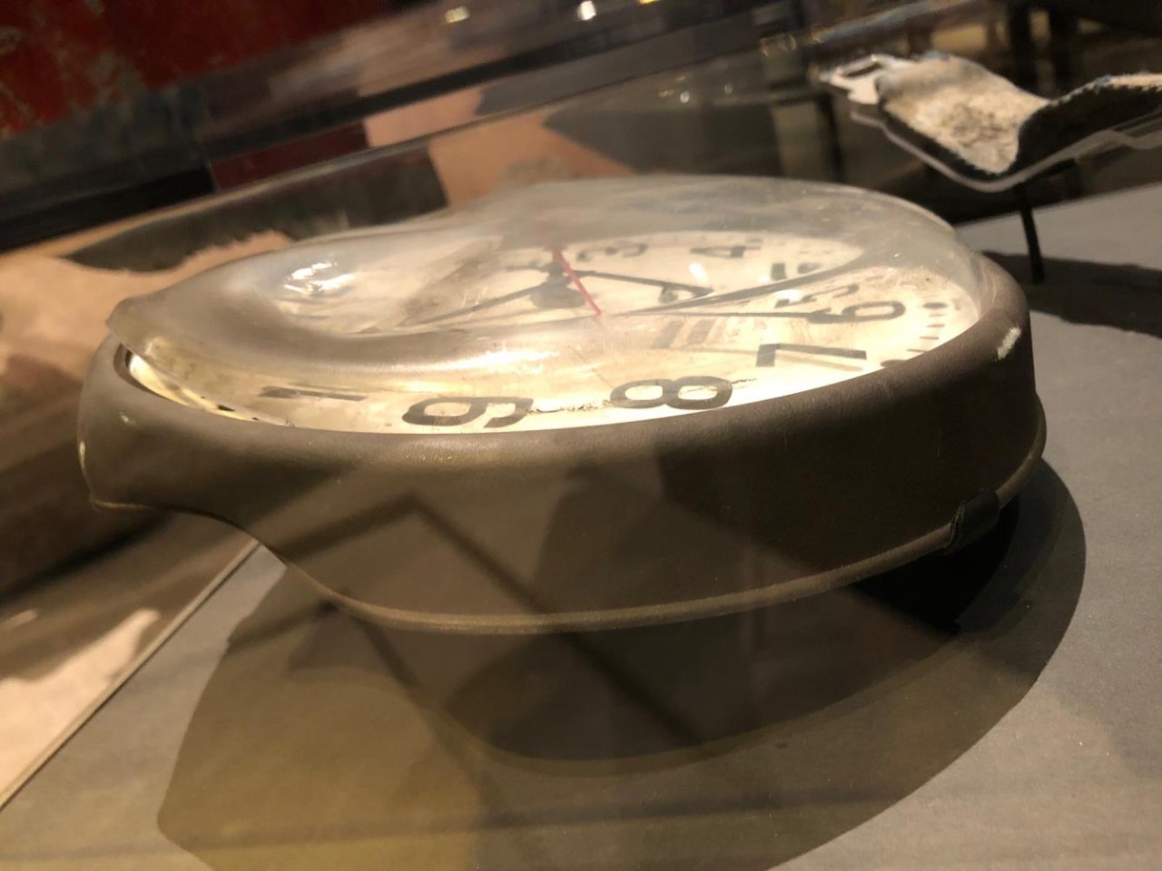 Profile view of 12-hour clock with white face and black numbers. Clear plastic cover blackened and warped from intense heat. Recovered from Pentagon after terrorist attack.