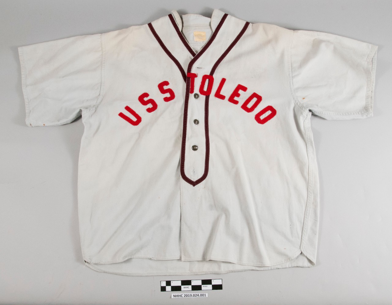 Obverse view of the baseball uniform blouse. Off-white in color with red lettering "USS Toledo." Two brown stripes around the collar of the blouse. Four brown plastic buttons on the obverse.