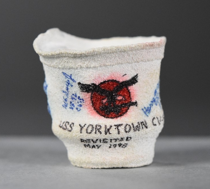 One crushed 8oz. Styrofoam coffee cup. On the obverse is a drawing of an eagle with outstretched wings, gripping a cannon in its talons, on a field of red (the insignia of USS Yorktown CV-5). Below the insignia is “USS Yorktown CV-5 / Revisited /...
