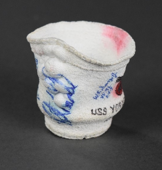 One crushed 8oz. Styrofoam coffee cup. It has a partial view of the USS Yorktown's insignia of an eagle carrying a cannon, the signature of Dr. Robert Ballard, and the signature of W.F. Surgi, VF-42, CV-5.