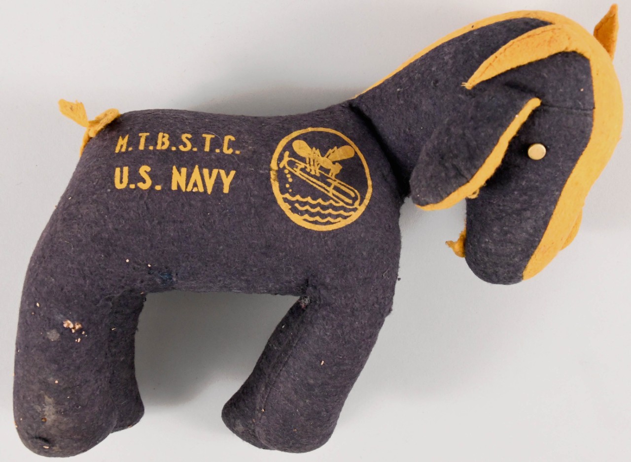 Stuffed blue felt goat mascot with gold mane and tail. M.T.B.S.T.C. U.S. Navy and PT mosquito logo prtined on its right side.